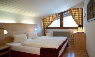 A cozy bedroom with a double bed and warm wooden tones. The windows allow plenty of daylight in, and the room is simply but stylishly furnished.