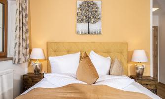 A cozy bedroom with a yellow headboard and soft pillows. A piece of artwork of a tree hangs on the wall, and there are two bedside lamps.