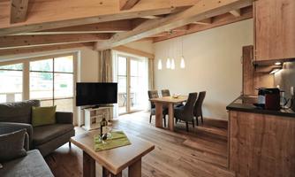 A modern living room with wooden flooring and large windows. The room features a cozy seating area, a dining table, and a kitchenette.