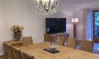 An elegant dining room with a large wooden table and comfortable chairs. Above the table hangs a beautiful chandelier and a television is mounted on the wall.