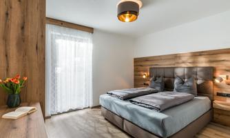 A modern bedroom with a large bed and wooden paneling. The windows let in plenty of daylight, and a fresh bouquet of flowers is on the table.
