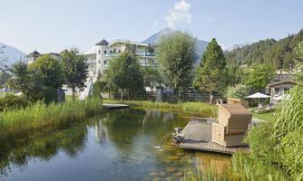 A quiet landscape with a pond and two wooden balconies. In the background, modern buildings and lush trees stand.