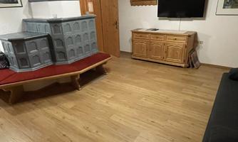A bright living room with a wooden floor. There is a couch, a dresser, a tiled stove, and a TV.