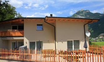 A modern house with wooden cladding and a cozy outdoor area. In the background, green mountains and a clear blue sky can be seen.