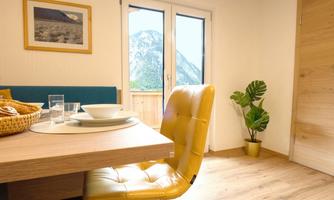 A cozy dining area with a yellow chair and a wood-colored table. A mountain view can be seen through the windows, and a plant stands in the corner.
