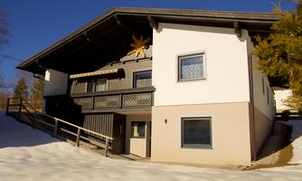 A modern wooden house with a slanted roof and a sunny facade. Snow surrounds the house, and there is a small staircase leading to the entrance.