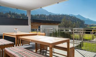 A sunny terrace with wooden furniture and a view of the mountains. The surroundings are green and inviting.