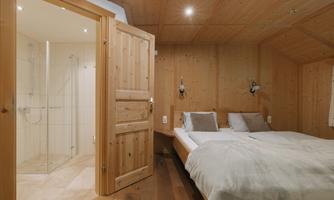 A cozy bedroom made of wood with a double bed and attached bathroom. The walls are clad in wood and there is pleasant lighting.