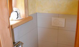 A small bathroom with a white toilet and wooden paneling. A painting is hanging on the wall, and on a shelf, there are towels and a washing utensil.