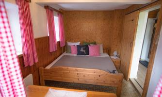 A cozy bedroom with a wooden bed and colorful cushions. Red checked curtains and warm wooden walls create an inviting atmosphere.