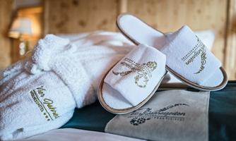 A fluffy bathrobe and slippers are lying on a bed. The textiles are white with golden lettering.