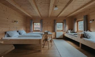 A cozy wooden room with two beds and a single bed. Large windows let in a lot of light and create an inviting atmosphere.