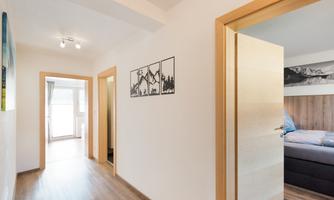 A bright hallway with wooden flooring and white walls. Pictures are hanging on the wall, and there is a door leading to another room.