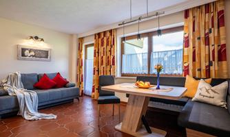 A cozy living area with a gray sofa, red cushions, and a wooden dining table. Large windows let in plenty of light, and the decorative curtains add color to the room.