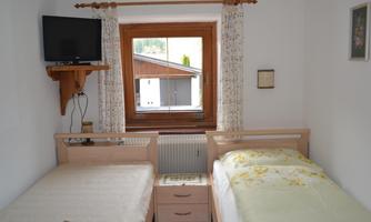 A cozy bedroom with two single beds and a TV on the wall. The window offers a view of the outside of the house.