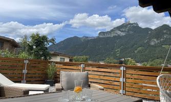 A cozy terrace with a view of the mountains. On the table are glasses and a magazine, surrounded by a natural environment.