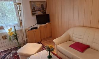 A cozy living room with a white sofa and a small table. Pictures hang on the wall, and a TV is placed on a wooden cabinet.