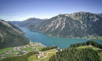 A picturesque landscape with a clear lake and majestic mountains. In the foreground, there are wooded hills and a small village.
