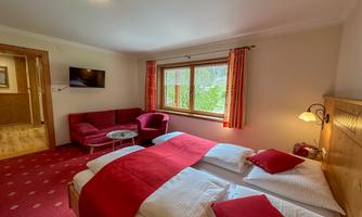 A cozy hotel room with double beds and red bedding. There is a television, a small seating area, and plenty of daylight through the windows.