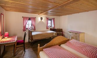 A cozy bedroom with two beds and warm wooden decor. The windows have red and white curtains, and there is a desk with a chair.