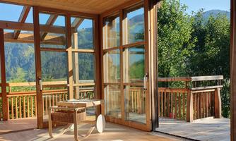 A bright, modern room with large wooden windows. The view leads to a green landscape and trees.