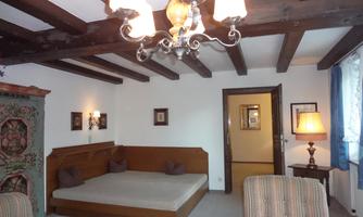 A cozy bedroom with a large bed and a wooden beam ceiling. The decor is simple and rustic, featuring a floor lamp and an antique wardrobe.