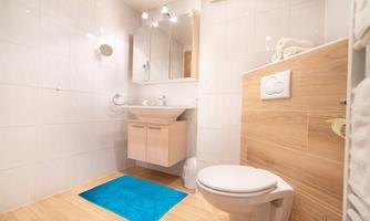 A modern bathroom with a bright tiled wall and wooden accents. There is a sink, a toilet, and a blue carpet on the floor.