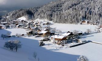 A winter landscape with a village covered in snow. In the background, there are snow-covered mountains and trees.
