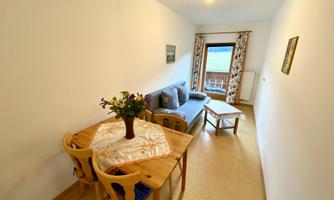 A living room with a cozy sofa and a dining table. The walls are light, and a window provides plenty of daylight.