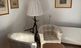 A cozy living room with two upholstered armchairs and a small table. Pictures hang on the wall, while a floor lamp provides warm light.