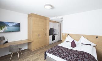 A modern room with a double bed, a television, and a small kitchenette. The floor is laid with wood and there are stylish furnishings.