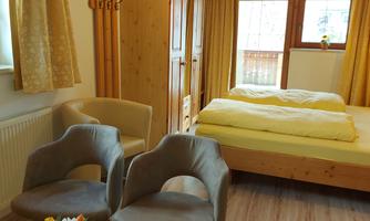 A cozy room with wooden furniture and a large window. There are two chairs, a table with flowers, and a double bed.