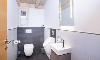 A modern bathroom with gray tiles and white sanitary fixtures. There is a window and a lit mirror.