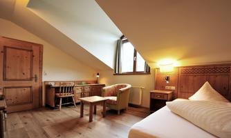 A cozy room with sloped walls and wooden furniture. There is a bed, a table, and a small seating area with a window that lets in daylight.