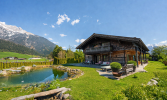 A cozy wooden house with a pond and a beautiful lawn. In the background, there are high mountains and a blue sky.
