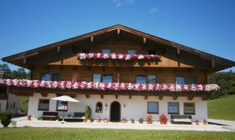 A traditional house in alpine style with colorful flowers at the windows. In the foreground, there is a small garden with seating and a sun umbrella.