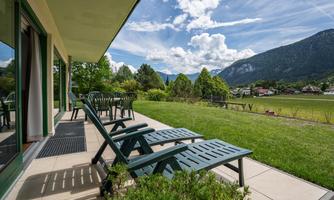 A terrace with loungers and a view of green meadows and mountains. The sky is clear and there are some clouds.