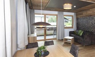A modern living room with wooden paneling and large windows. The room is cozily furnished with a dining table, chairs, and a couch.