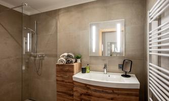 A modern bathroom with a spacious shower and a stylish wooden dresser. The mirror is equipped with LED lighting and towels are available.