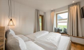 A cozy bedroom with two beds and light wood. Large windows with curtains offer a view outside.