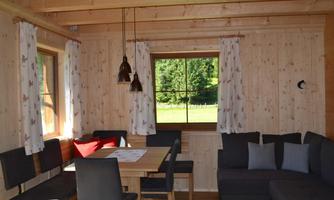 A cozy living space with wooden walls and a large window. There is a dining table, chairs, and a comfortable couch.