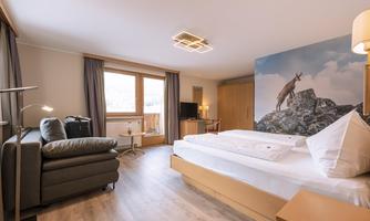 A modern hotel room with a large bed and a cozy sofa. On the wall, there is a picture of a chamois, and the windows provide daylight.
