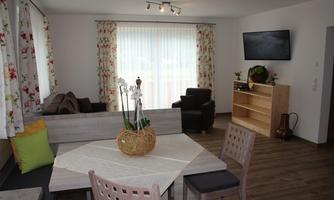 A modern living room with a dining table, cozy seating, and a television. Large windows let in plenty of daylight, and the floral decoration creates an inviting atmosphere.