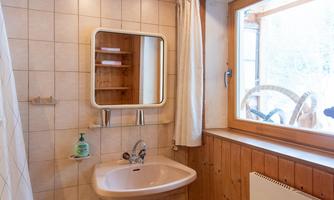 A simple bathroom with a sink, a mirror, and a window view. The walls are partially tiled and made of wood.