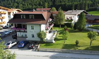 A charming guesthouse with a green garden and blooming plants. In the foreground, cars are parked and in the background, mountains are visible.