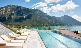 A modern swimming pool with lounges, surrounded by impressive mountains and a clear lake. The sky is blue and cloudless, creating a relaxing atmosphere.