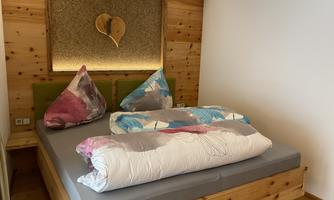A cozy bedroom with a large bed and colorful cushions. The wall is designed with wood and there is warm light.