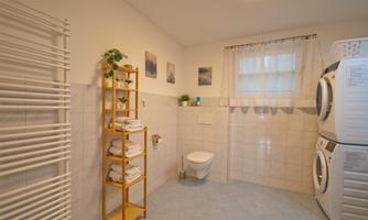A bright, well-furnished bathroom with a washing machine and dryer. A shelf with towels and decorative plants completes the space.