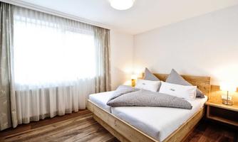 A bright bedroom with a large double bed and cozy pillows. The room has large windows and a warm wooden floor.