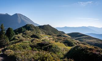 A picturesque mountain landscape with gentle hills and green meadows. In the background, majestic mountains can be seen under a clear blue sky.
